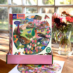 Jigsaw puzzle box with a colorful design of birds and flowers on a wooden table with a vase of flowers in the background.
